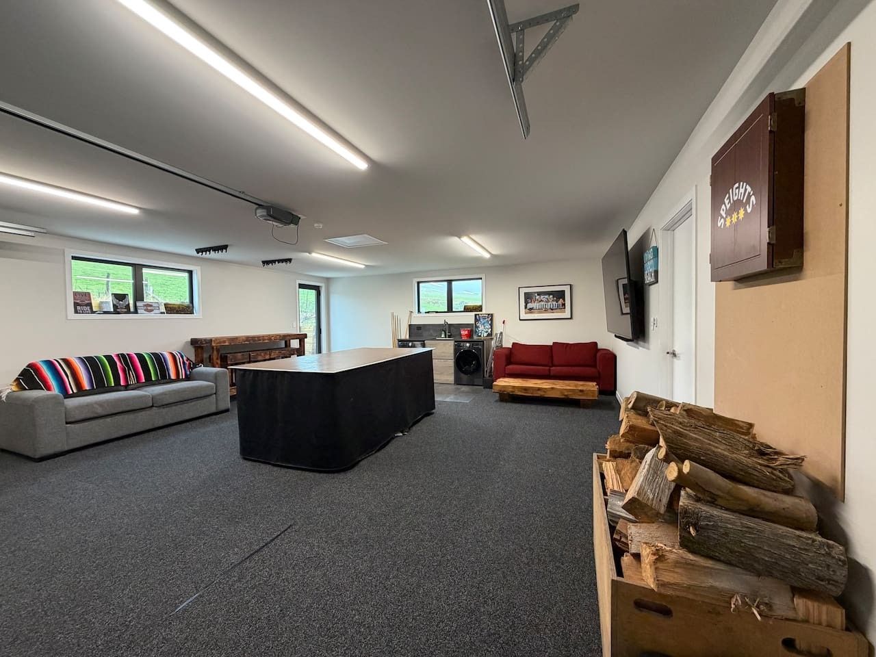 A spacious room with a large black table, a grey sofa, a red sofa, and a wooden bench. There are windows with views of greenery, a fireplace, and a wall-mounted shelf with a sign.