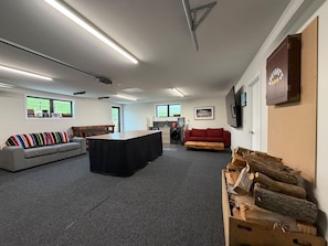 A spacious room with a large black table, a grey sofa, a red sofa, and a wooden bench. There are windows with views of greenery, a fireplace, and a wall-mounted shelf with a sign. - Luxe Lookout Riverton (Riverton / Aparima)