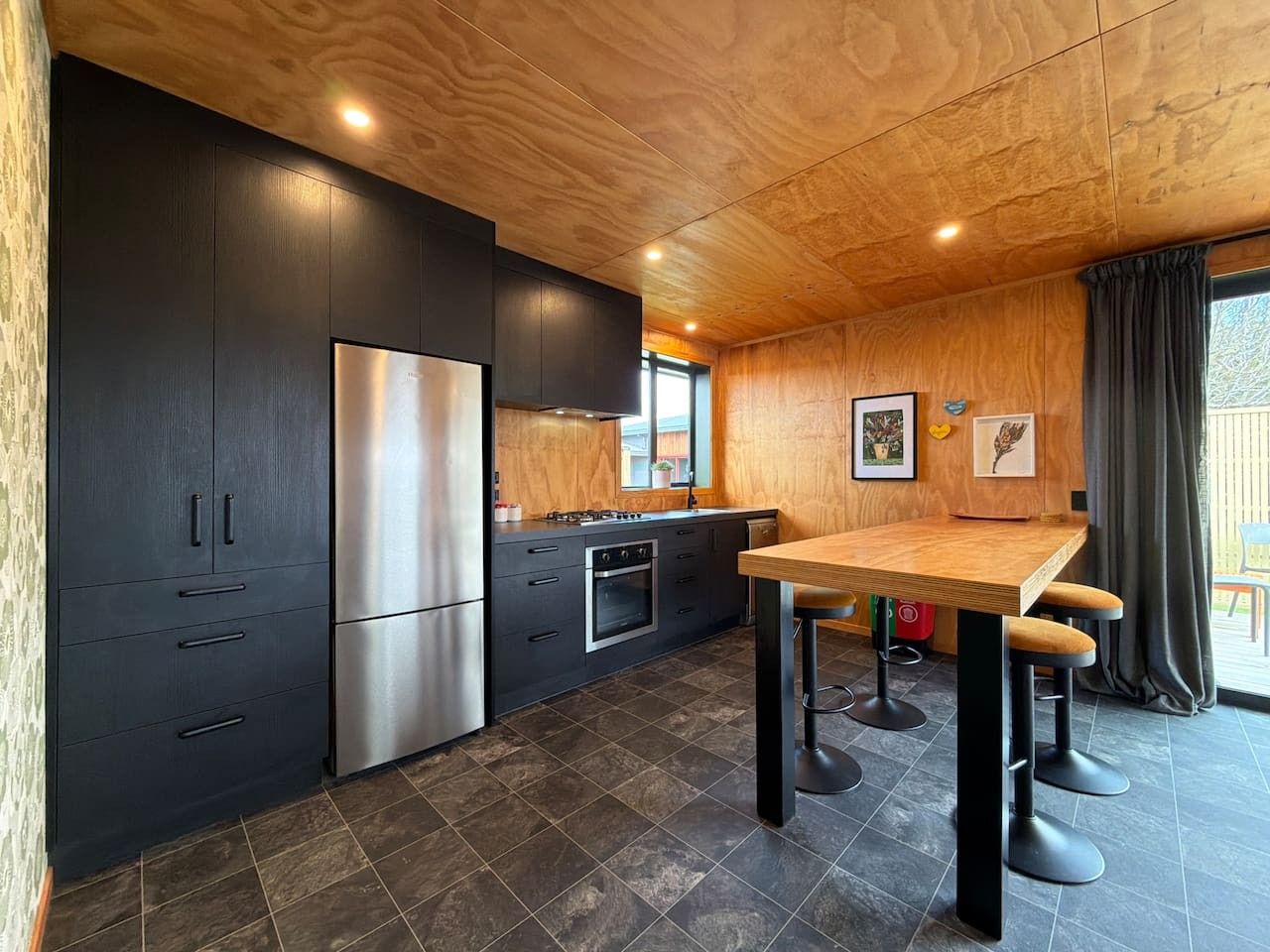 A modern kitchen with dark cabinetry, stainless steel appliances, and a wooden dining table with bar stools.