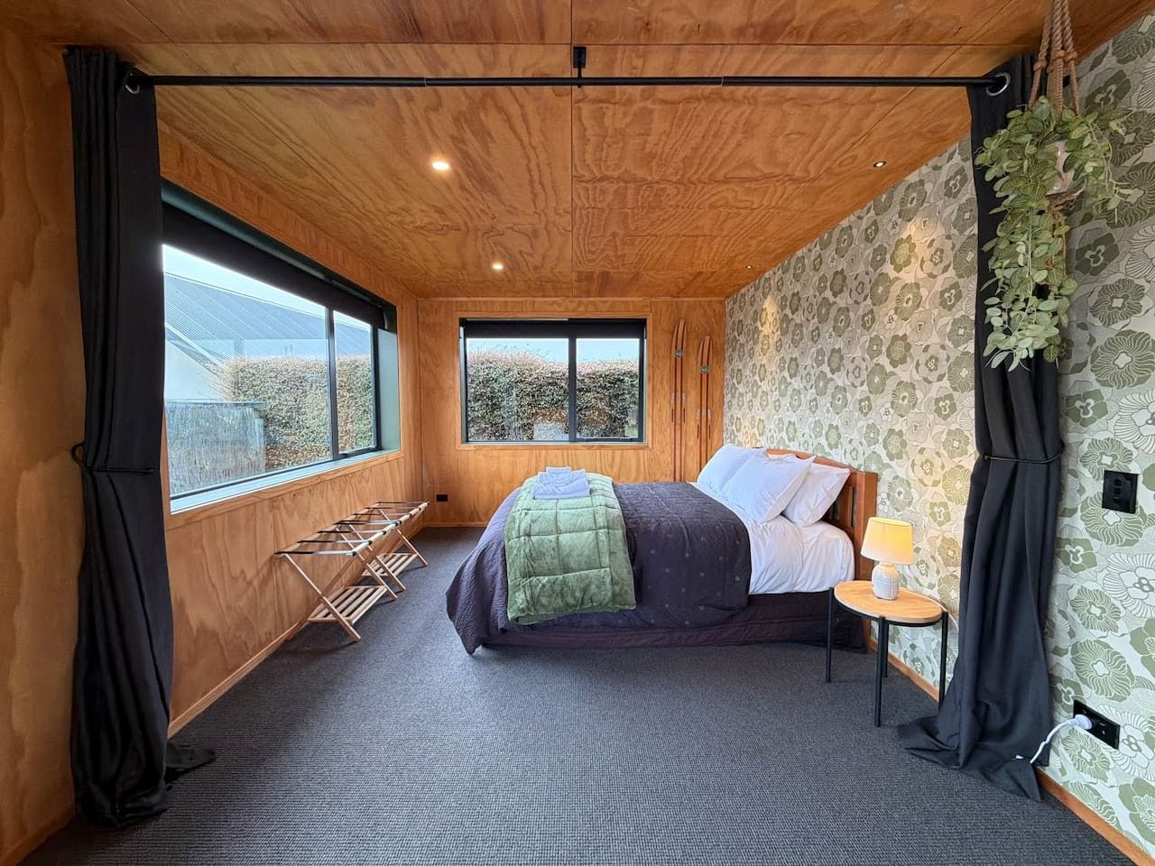 A room with a bed, a window, a hanging plant, and a small table with a lamp.