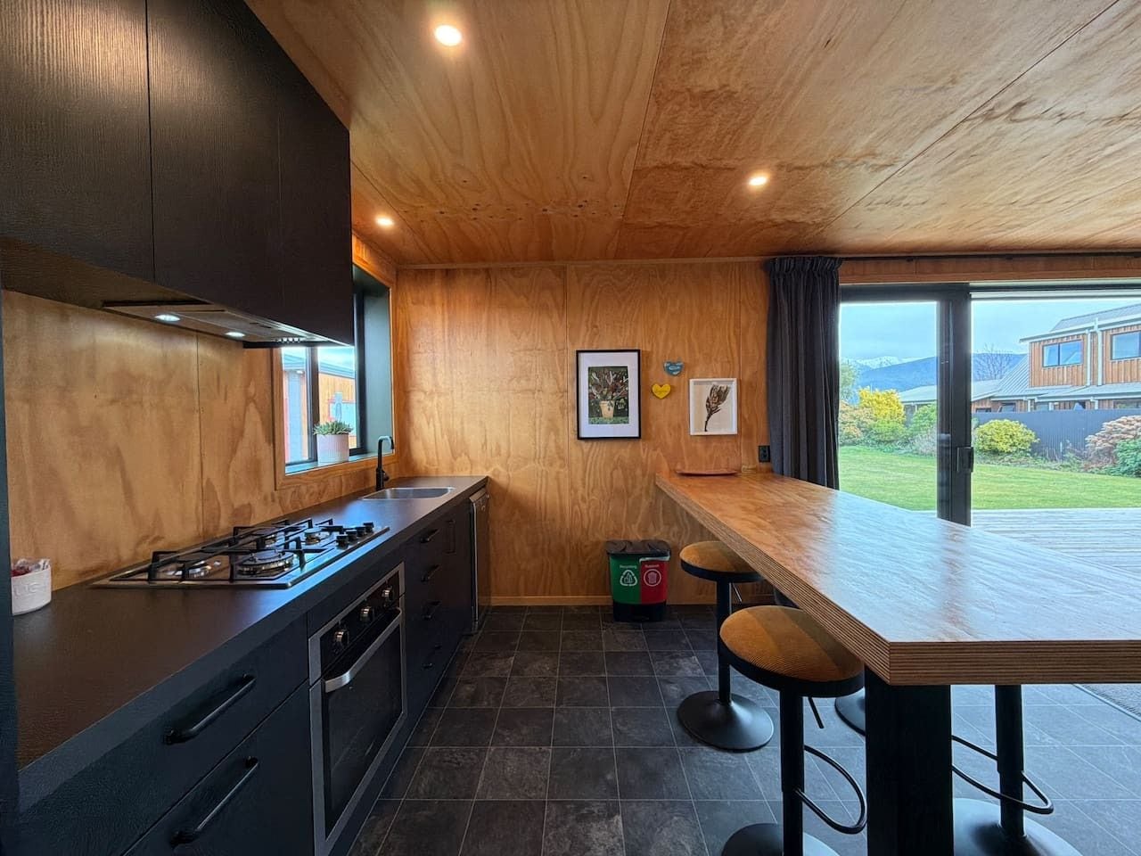 Une cuisine moderne avec un plafond en bois, une cuisinière à gaz, des meubles de rangement aux couleurs sombres et une grande fenêtre donnant sur un jardin.