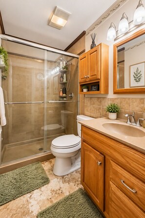 A bathroom with a glass shower enclosure, a toilet, wooden cabinets, a sink, and a mirror. - Overland Park Retreat with Pool & Shuffleboard! (Overland Park)