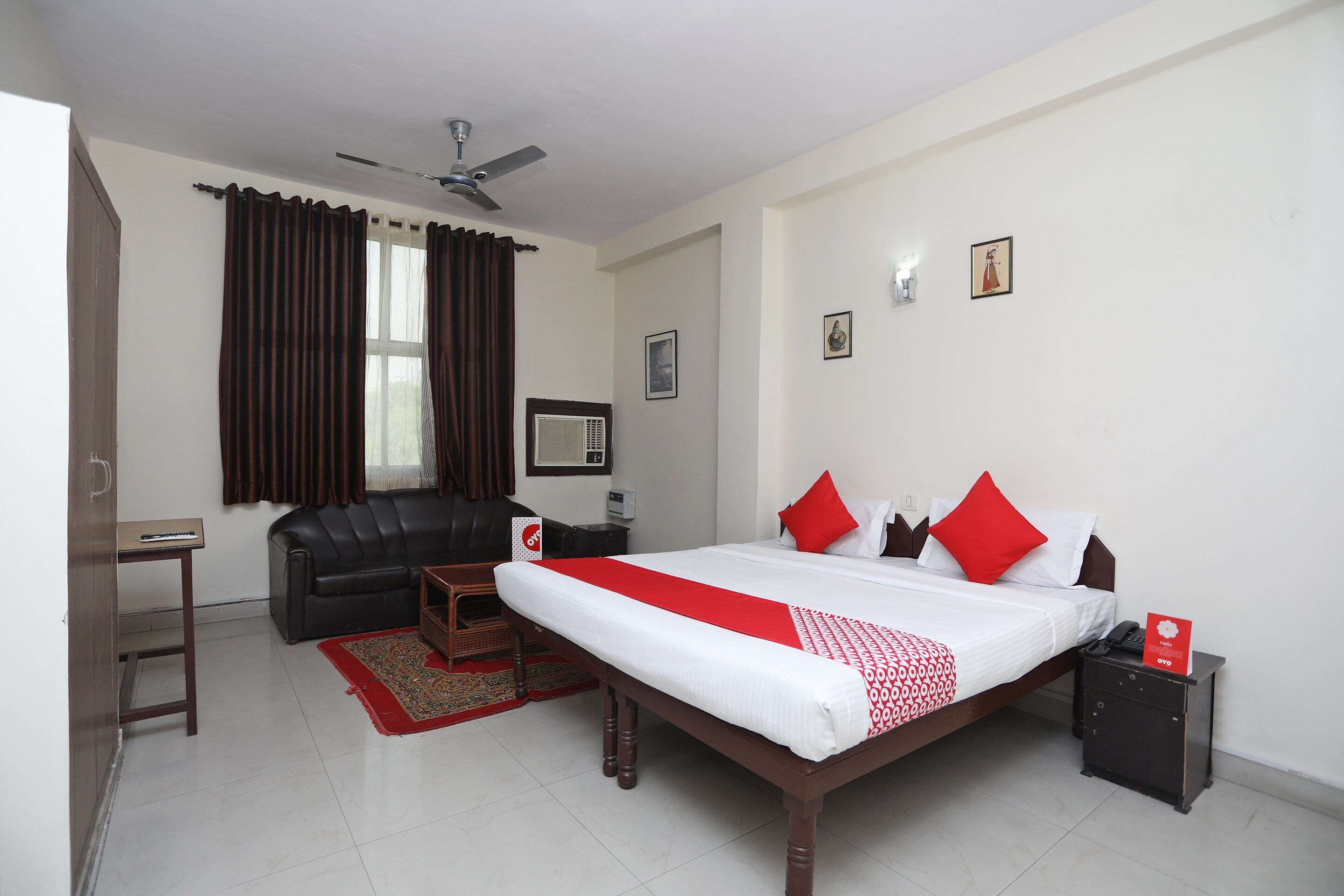 A hotel room with a bed, a desk, a chair, a sofa, a ceiling fan, and a window with curtains.