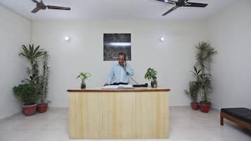 A reception desk with a man standing behind it, two ceiling fans, and potted plants on either side.