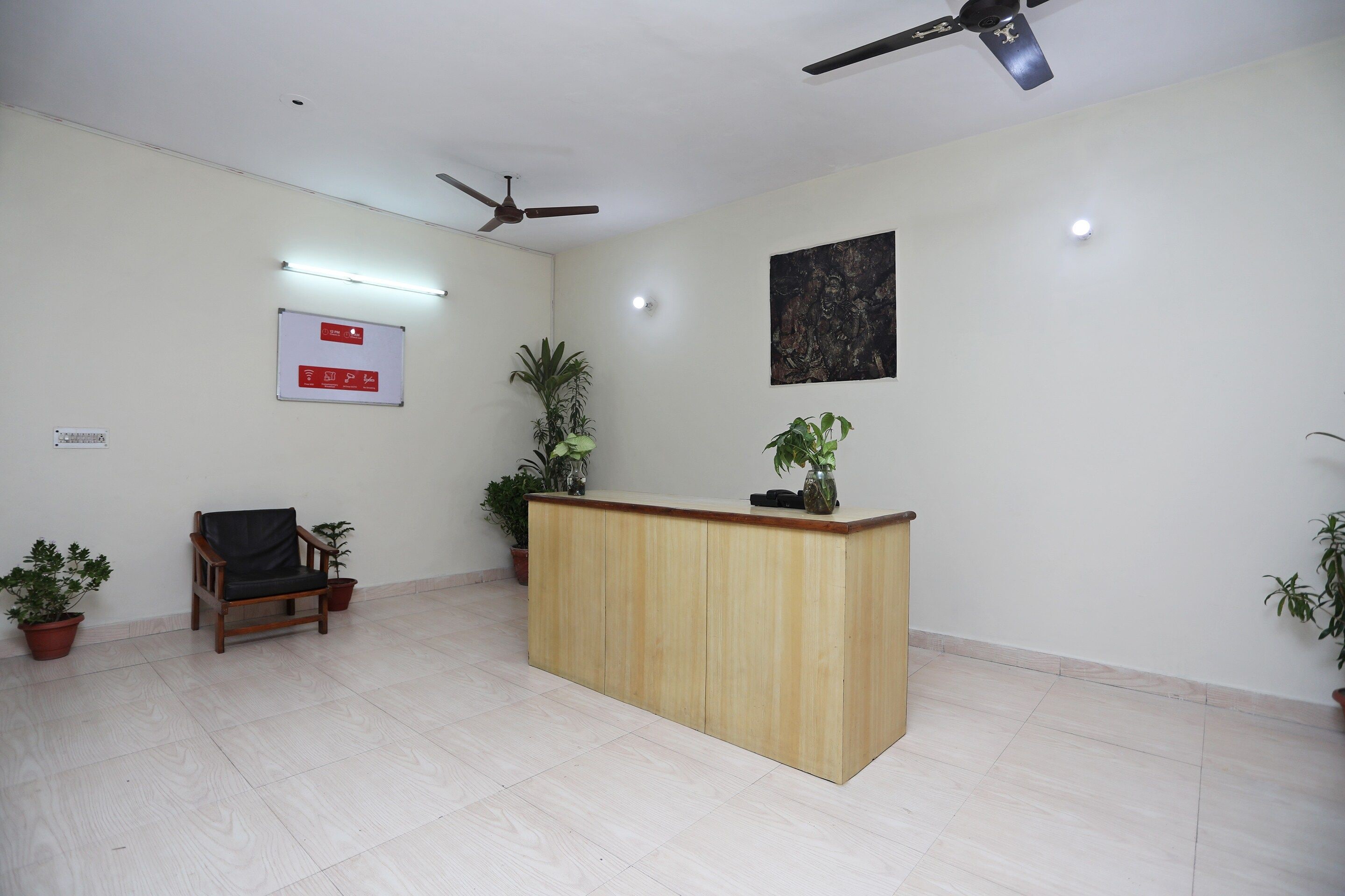 A reception area with a wooden desk, a black chair, potted plants, a ceiling fan, and a wall-mounted sign.