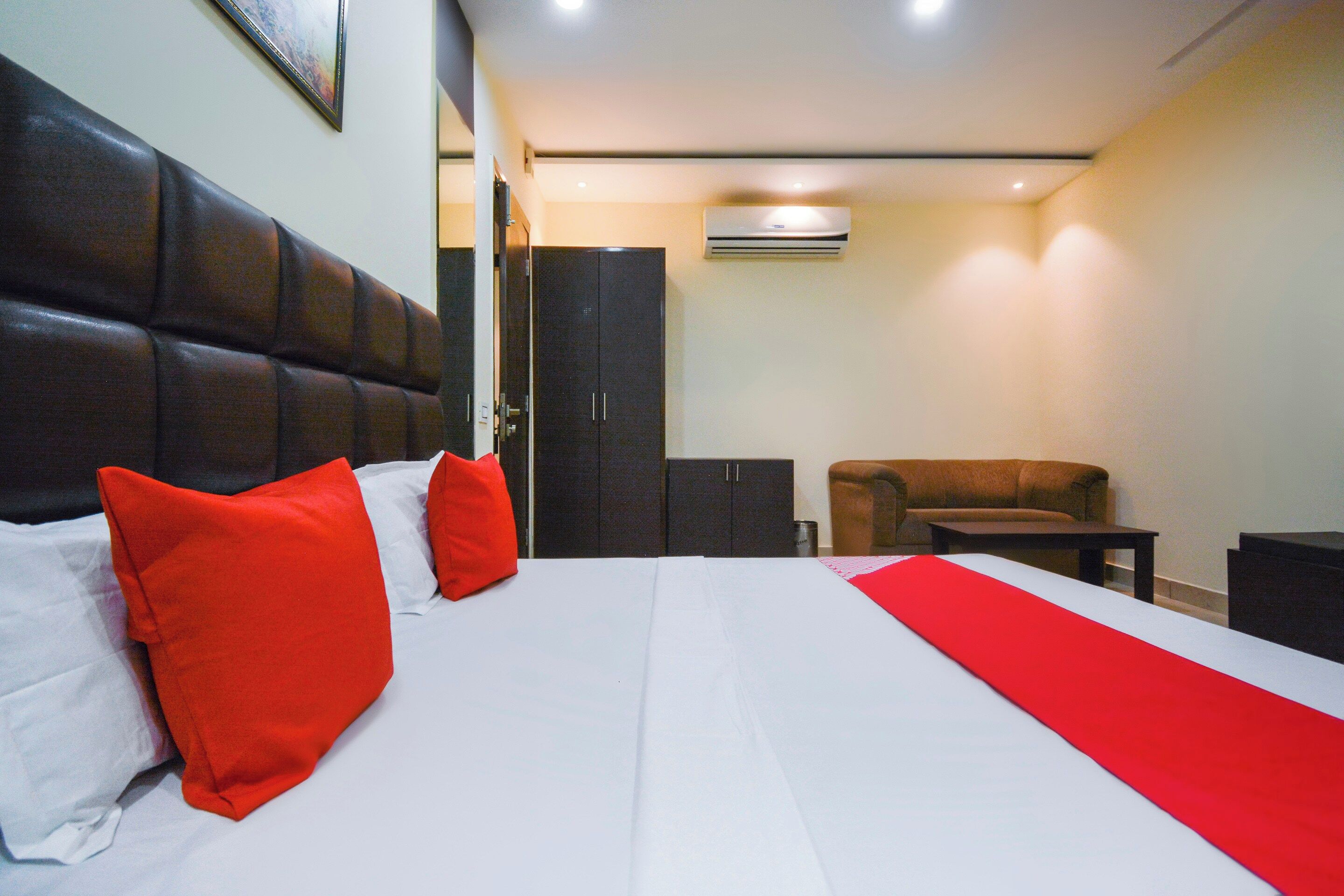Une chambre d’hôtel avec un grand lit, un plaid rouge, deux coussins rouges, une tête de lit en cuir marron, un canapé marron, une petite table et une armoire.