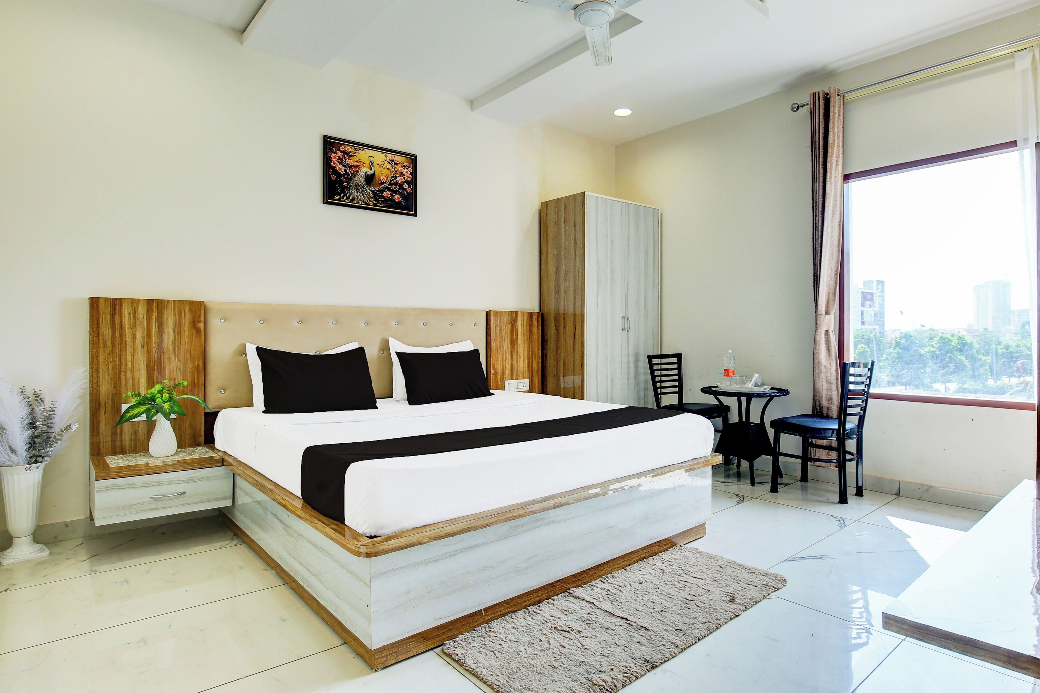 A modern hotel room with a large bed, a bedside table, a small table with chairs, a wardrobe, and a window with a view.
