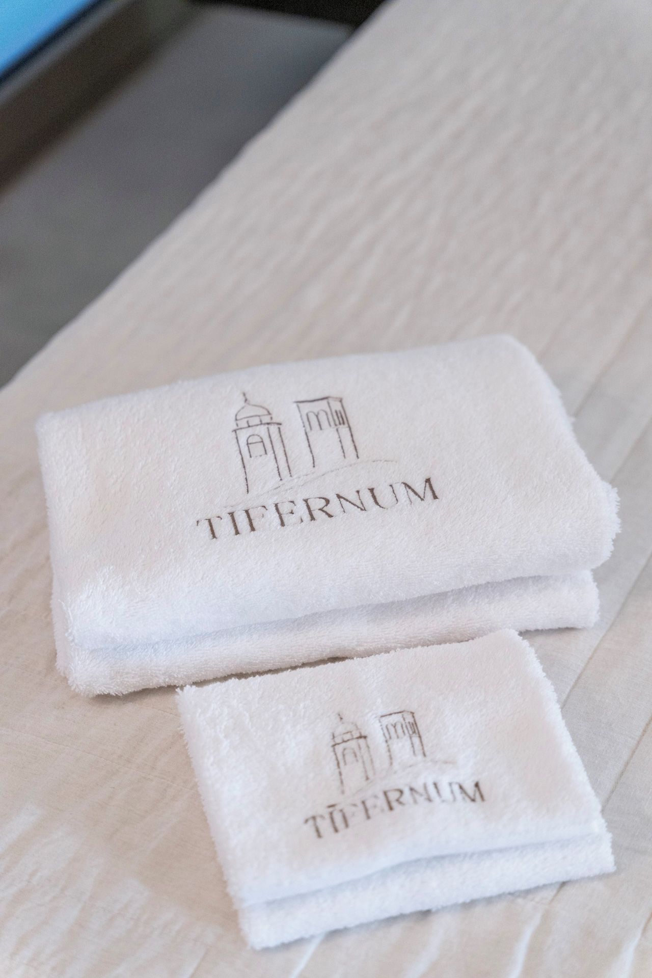 Deux serviettes blanches pliées, portant l’inscription « TIFERNUM » et une illustration représentant un bâtiment, sont posées sur une surface en bois.