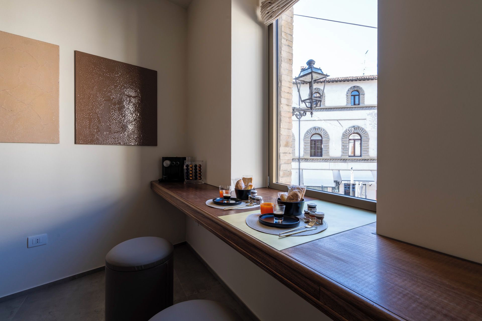 Un coin petit-déjeuner avec une machine à café, du pain et du jus, installé sur un comptoir en bois près d’une fenêtre donnant sur des immeubles.