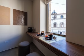 Un esmorzar preparat amb una màquina de cafè, pa i suc de fruita damunt un taulell de fusta a prop d'una finestra amb vistes a edificis.