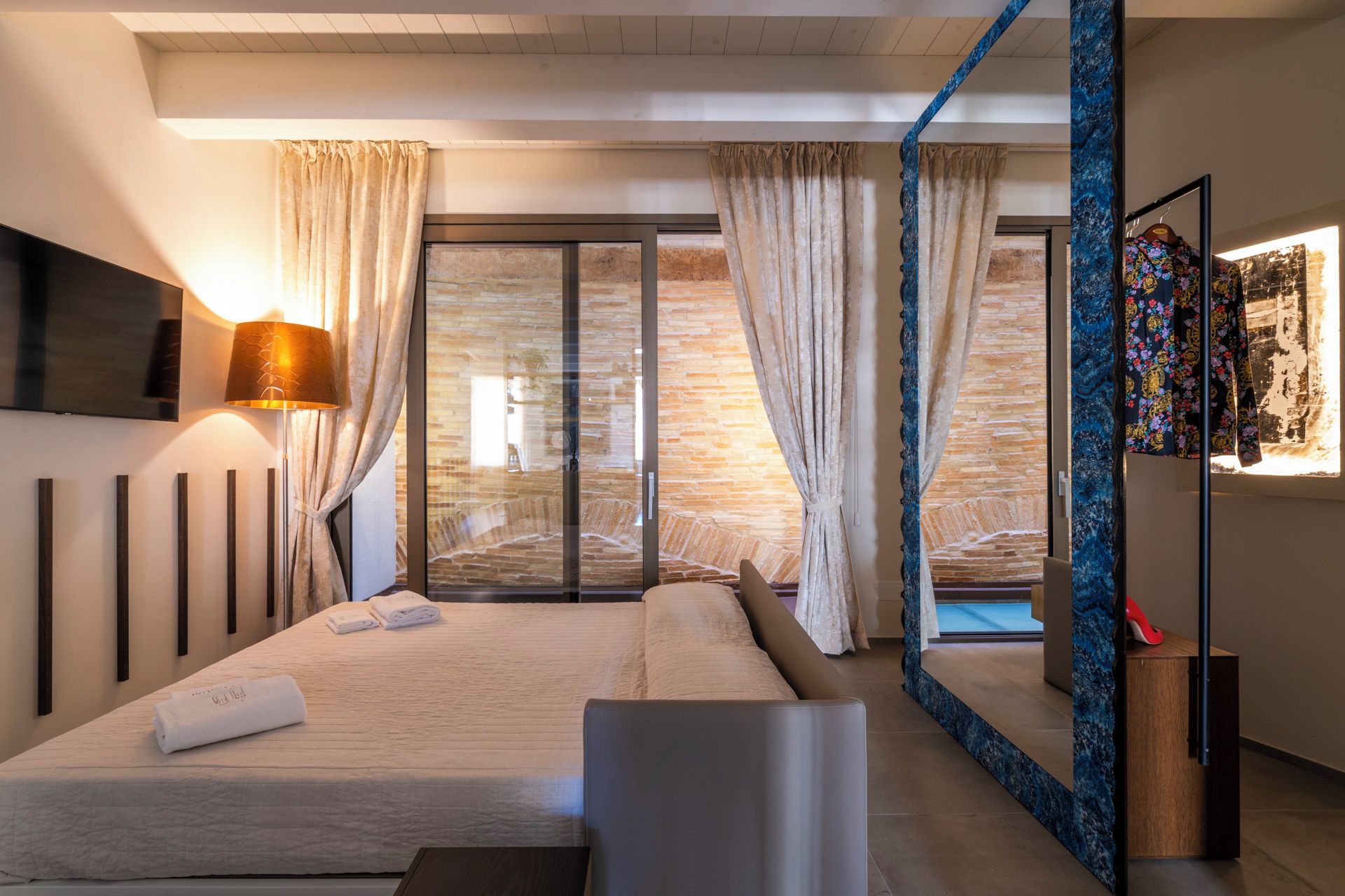 Une chambre moderne avec un grand lit, un téléviseur à écran plat fixé au mur, une lampe sur pied et une vue sur un mur en pierre, visible à travers une porte vitrée.