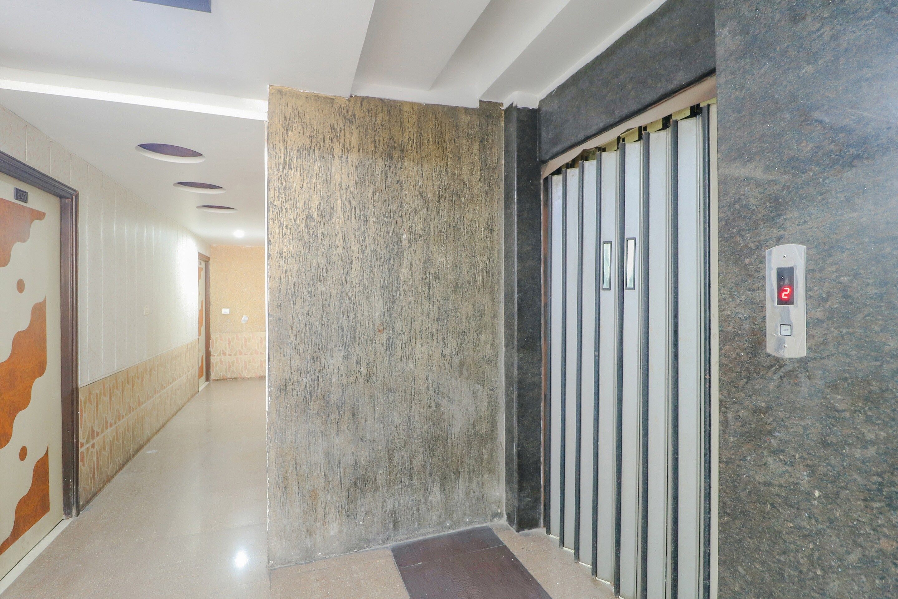 Un couloir avec un mur à texture, une porte dotée d’une poignée verticale et un panneau de commande numérique pour l’ascenseur.