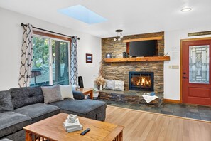 A living room with a stone fireplace, a large window with curtains, a sofa, and a flat-screen TV mounted above the fireplace. - Hot Tub + Pine-View Decks • Near Speedboat Beach + Crystal Bay | Emberpine (Kings Beach)