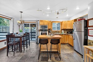 A modern kitchen with wooden cabinets, stainless steel appliances, a central island with bar stools, and a dining area with a table and chairs. - Hot Tub + Pine-View Decks • Near Speedboat Beach + Crystal Bay | Emberpine (Kings Beach)