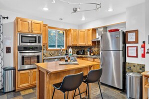 A modern kitchen with wooden cabinets, stainless steel appliances, a central island with chairs, and a tiled backsplash. - Hot Tub + Pine-View Decks • Near Speedboat Beach + Crystal Bay | Emberpine (Kings Beach)