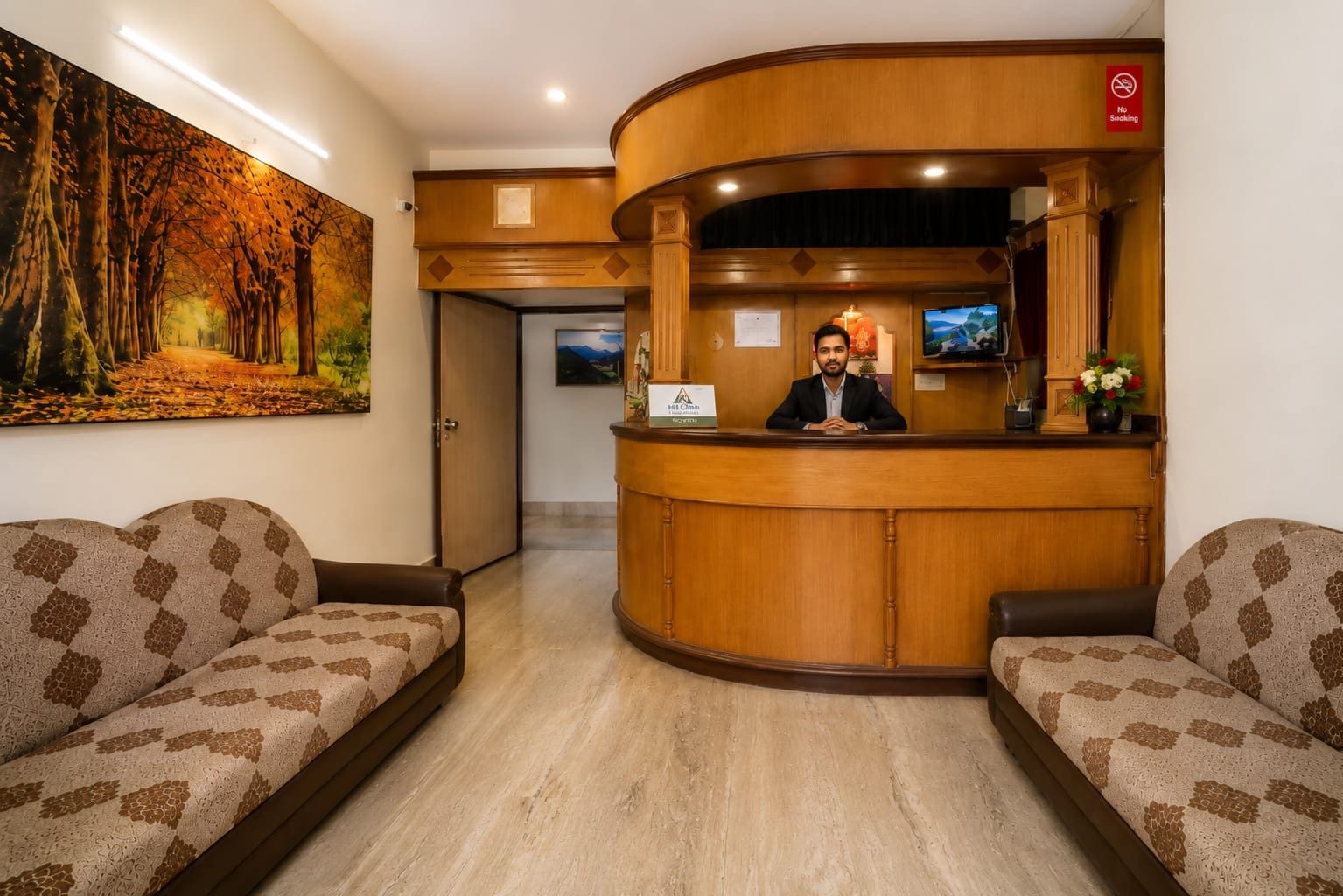 Un espace d’accueil avec un comptoir de réception en bois, deux canapés à motifs, un tableau représentant une forêt et un panneau « Il est interdit de fumer ».