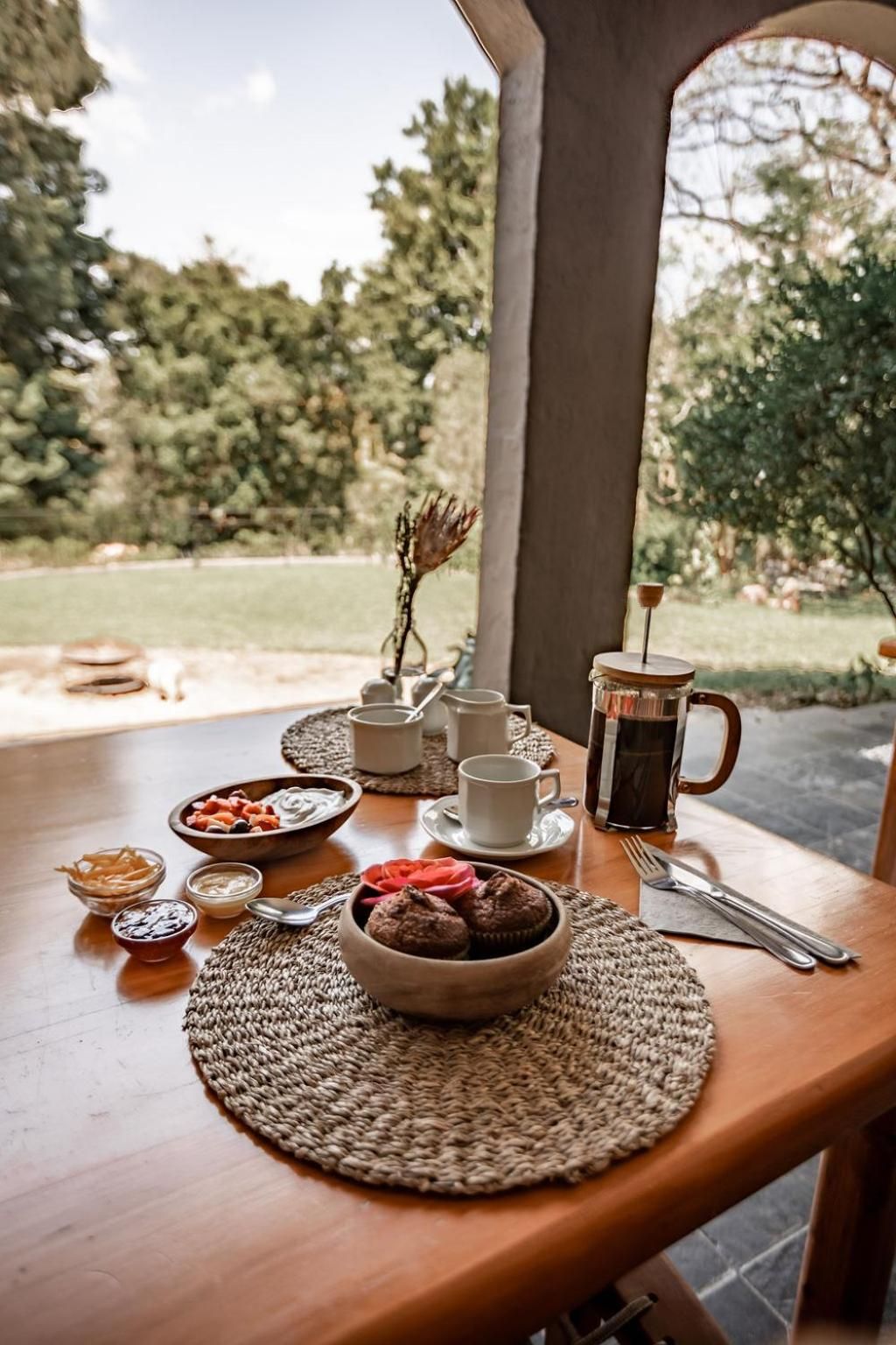 A wooden table set with a coffee pot, cups, a bowl of fruit, and a woven placemat.