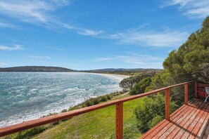 Eine Holzterrasse mit Blick auf einen Strand und das Meer, umgeben von Grünflächen und Hügeln in der Ferne.