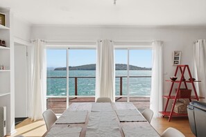 Ein Zimmer mit großem Fenster und Blick auf ein Gewässer, einem Esstisch mit Stühlen und einem roten Regal mit Körben.