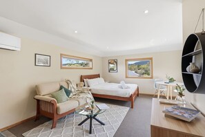 A modern bedroom with a bed, a sofa, a glass coffee table, and a desk with a chair. There are framed pictures on the wall and a view of the outdoors through the windows. - Cosy Bush Studio Near Airport (Acton Park)