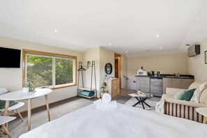 A modern hotel room with a bed, a dining table, a sofa, a kitchen area, and a large window with a view of greenery. - Cosy Bush Studio Near Airport (Acton Park)