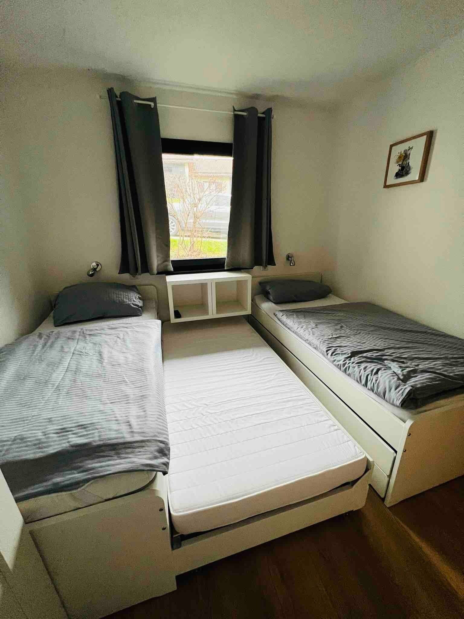 Ein kleines, kompaktes Zimmer mit zwei Einzelbetten, einem Fenster mit Vorhängen und einer kleinen Regalwand.