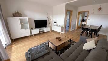 Una sala moderna con un sofá gris, un televisor sobre un soporte blanco, una zona de comedor con mesa y sillas, y una cocina al fondo.