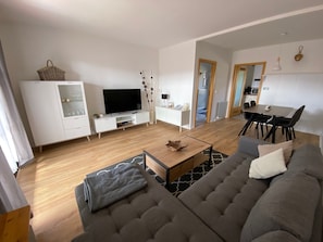 A modern living room with a grey sofa, a TV on a white stand, a dining area with a table and chairs, and a kitchen in the background.