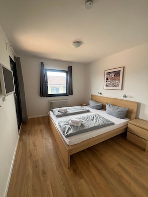 Ein Schlafzimmer mit einem hölzernen Bett, einem Nachttisch, einem Fenster mit Vorhängen, einem Heizkörper und einem gerahmten Bild an der Wand.