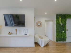 Ein modernes Wohnzimmer mit einem Flachbildfernseher, einer weißen Wand mit dekorativem Spiegel, Holzboden und einer grünen Wand mit Pflanzen.