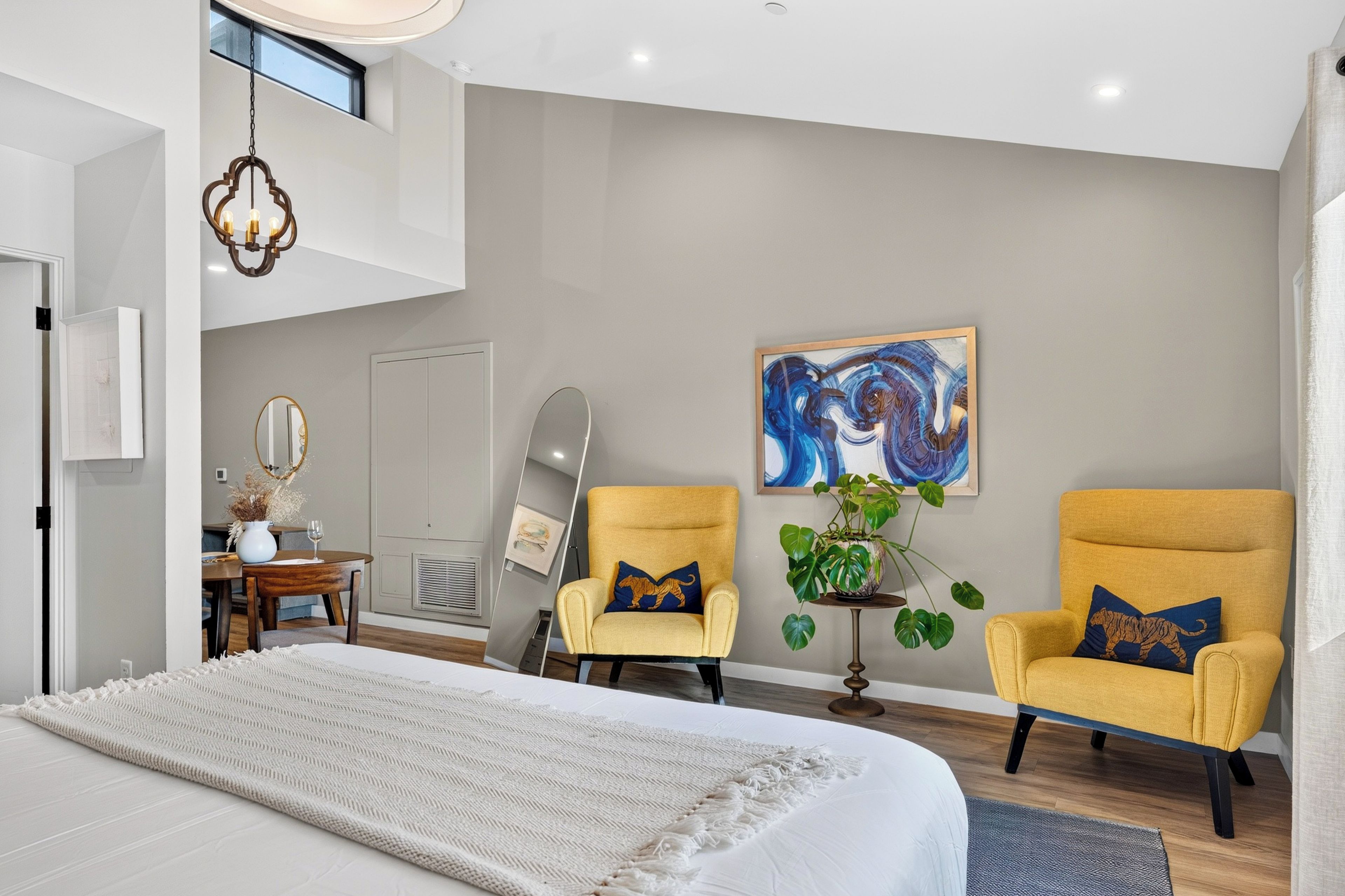 A modern bedroom with a large bed, two armchairs, a dining table, and a painting on the wall.