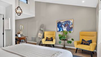 A modern bedroom with a large bed, two armchairs, a dining table, and a painting on the wall.