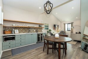 A modern kitchen with a dining area, featuring a round wooden table, light blue cabinets, and a decorative chandelier. - Paso Market Walk + Coffee + Dining | Mins to River Oaks | The Rosewood Room (Paso Robles)