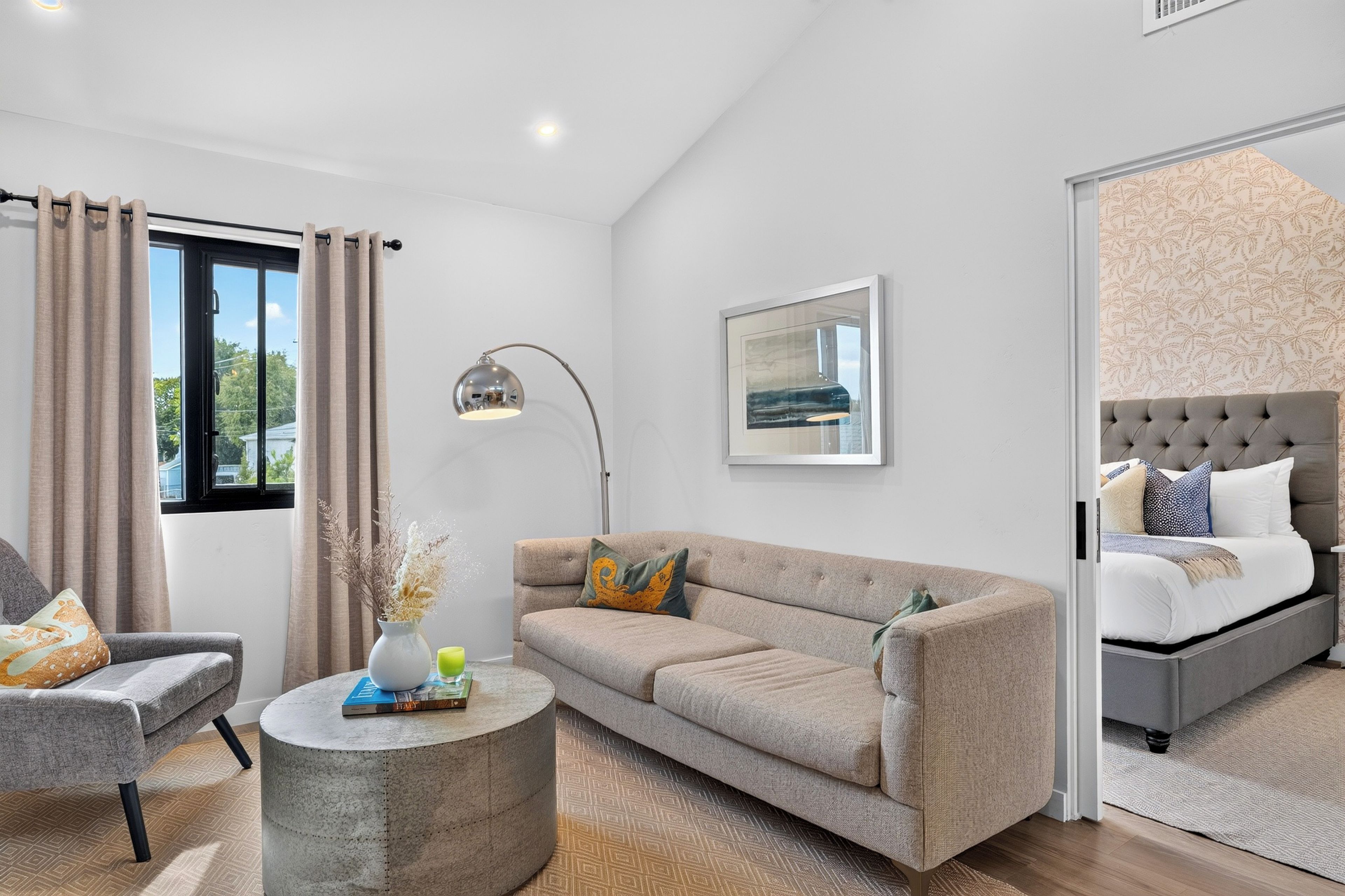 A modern bedroom with a grey sofa, a round coffee table with books, a bed with a grey headboard, and a large window with curtains.