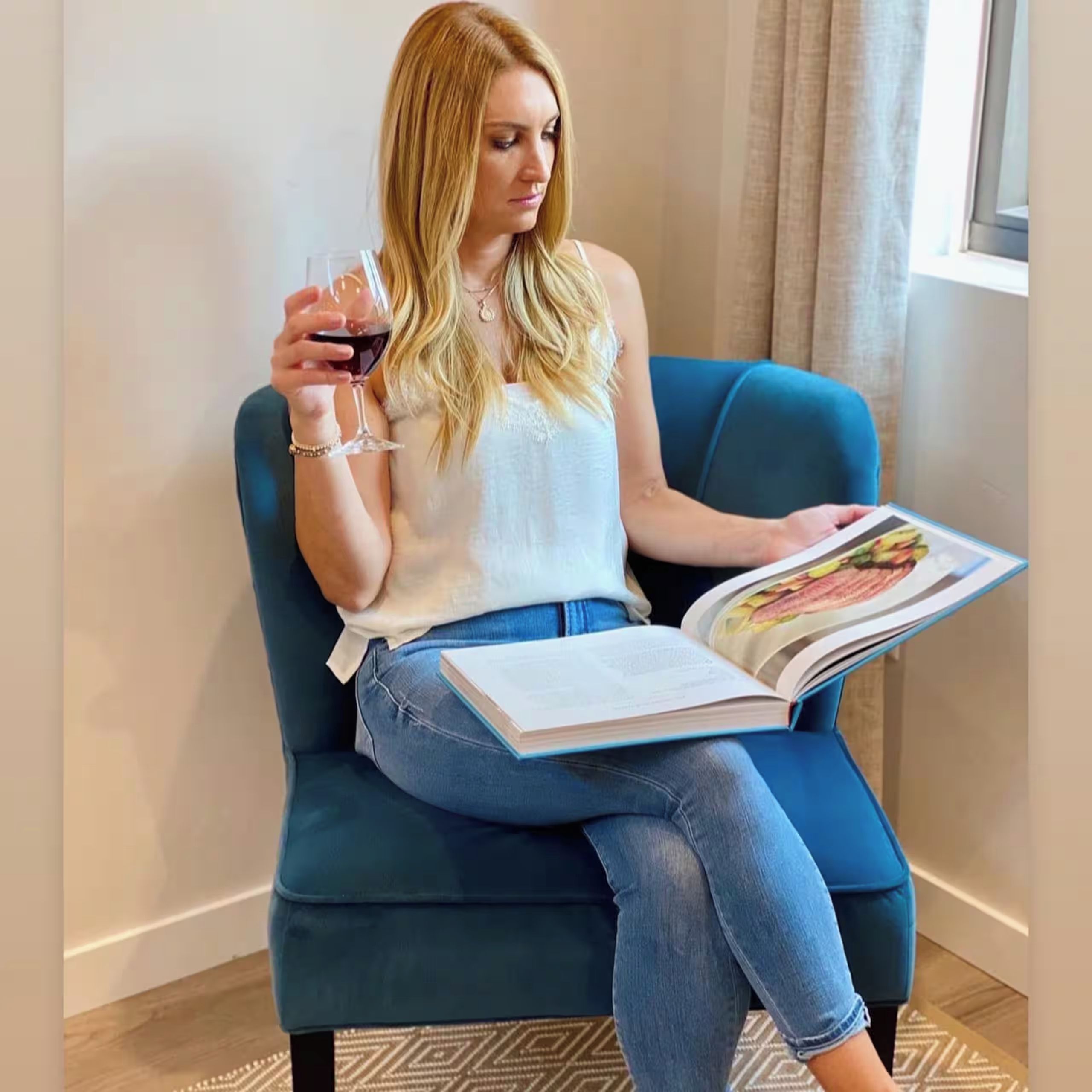 Una persona sentada en un sillón azul, sosteniendo una copa de vino tinto y leyendo un libro.