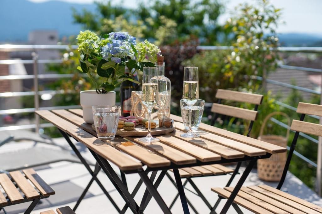 Une table en bois, agrémentée d’un pot de fleurs, de verres et de bouteilles, est placée sur une terrasse extérieure offrant une vue agréable.
