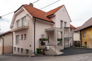 Um edifício residencial de dois andares com varanda, escadaria e um pequeno jardim frontal.