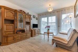 Een woonkamer met een houten kast, een televisie, een bank, een kleine tafel met een vaas en een groot raam met gordijnen.