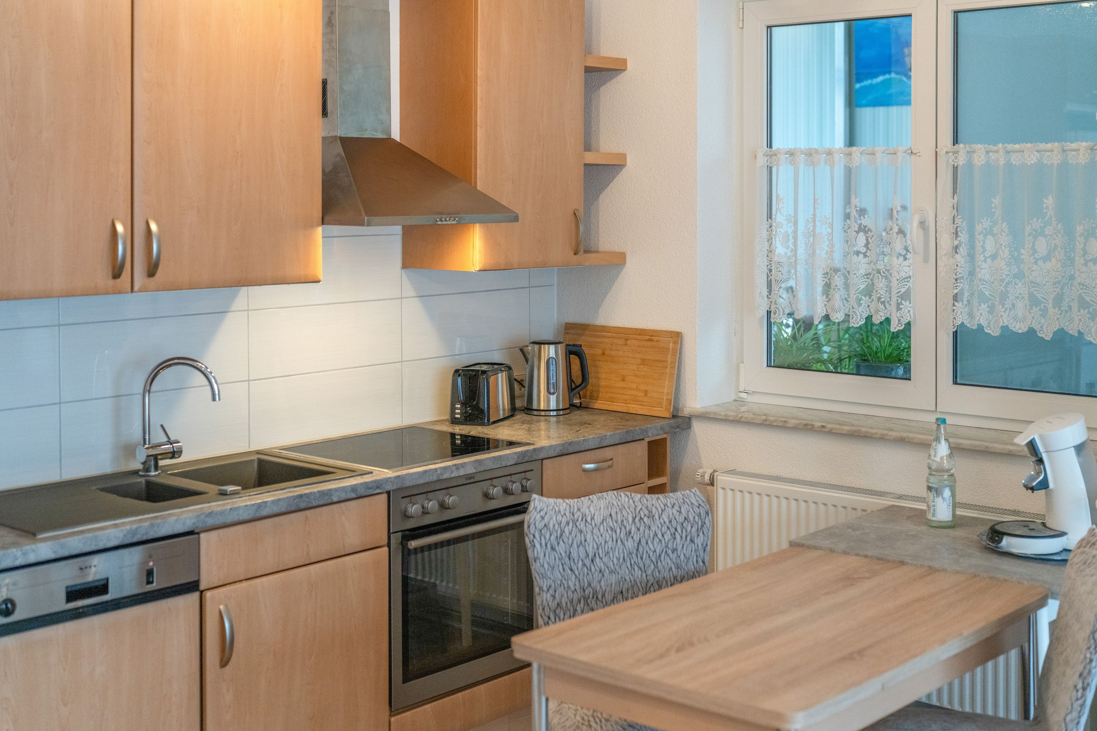 Une cuisine avec des meubles en bois, un évier en acier inoxydable, une cuisinière et une fenêtre ornée de rideaux en dentelle.