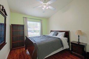 A bedroom with a bed, a nightstand, a lamp, a window with blinds, a ceiling fan, and a wooden wardrobe. - Travel Nurses Welcome! Zephyrhills Extended Stay (Zephyrhills)