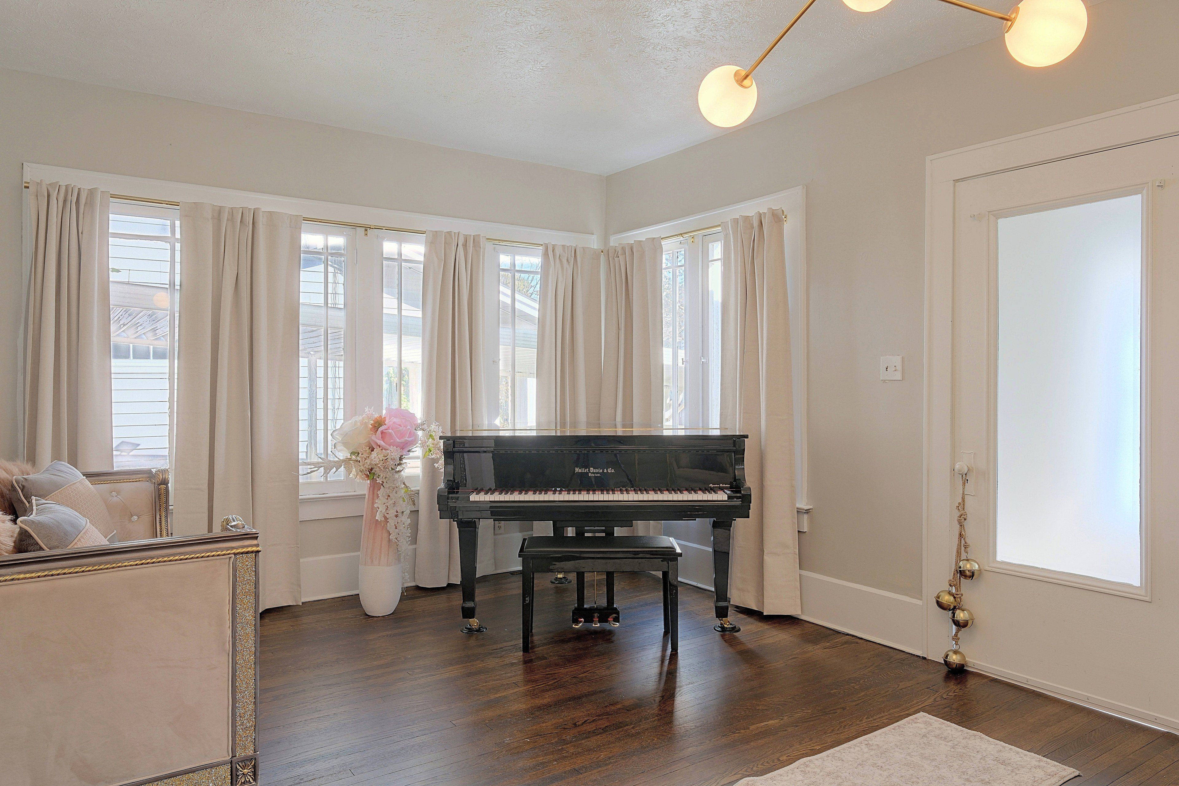 Une pièce spacieuse avec un grand piano, de grandes fenêtres avec des rideaux, une porte blanche et un plancher en bois.