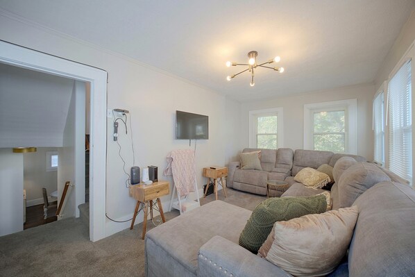 A living room with a sectional sofa, a flat-screen TV mounted on the wall, and a ceiling light fixture.