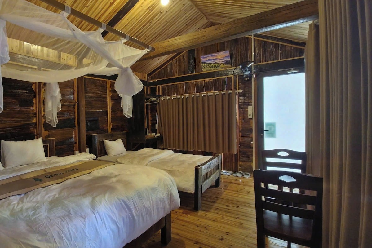A wooden cabin room with two beds, a wooden ceiling, and a large window with curtains.