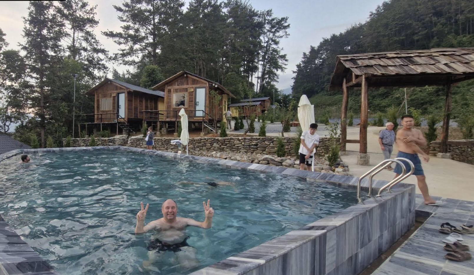 Una piscina con borde de piedra, terraza de madera y un área cubierta con techo metálico.