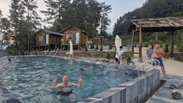 Una piscina con borde de piedra, terraza de madera y un área cubierta con techo metálico.
