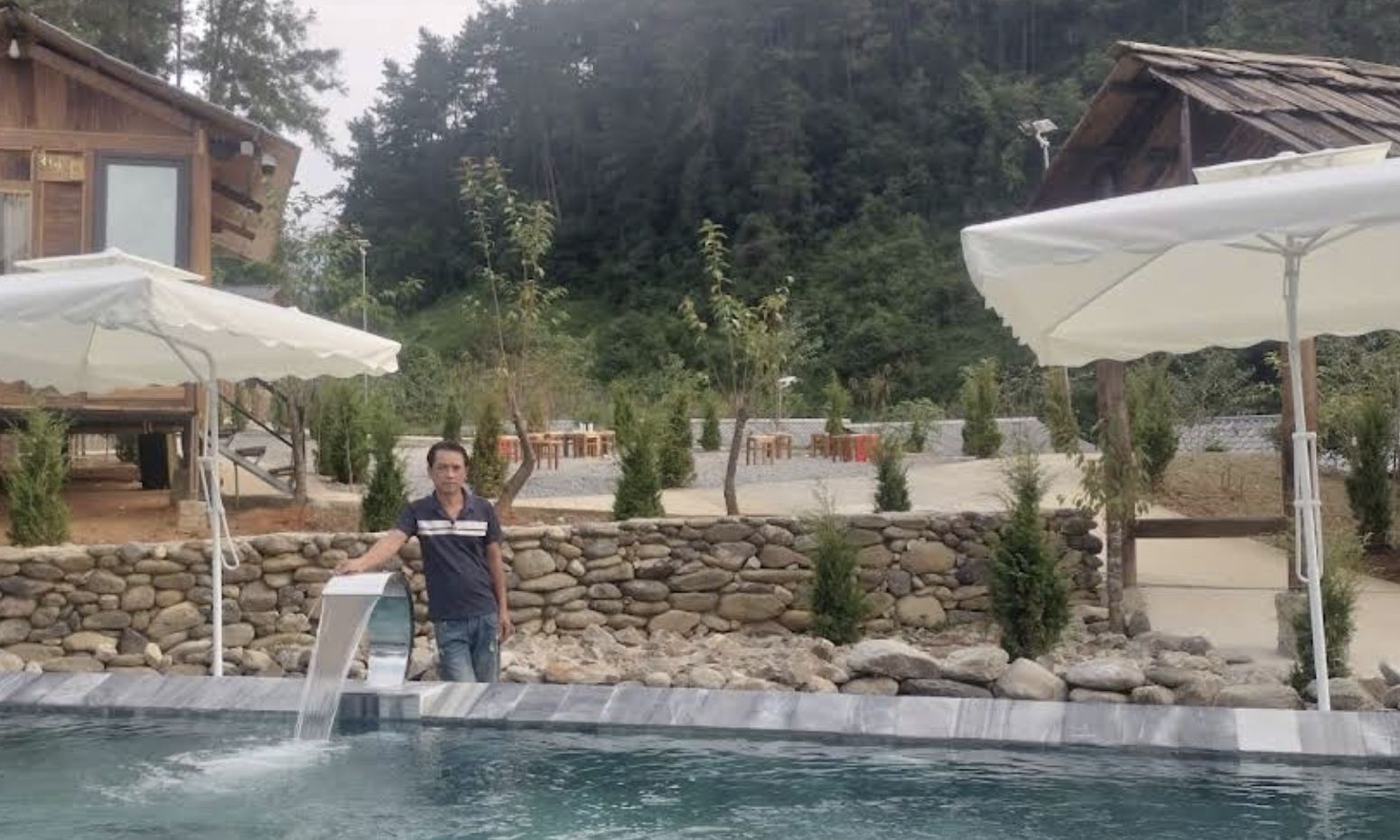Zona de piscina amb un mur de pedra, una persona al costat de la piscina i un edifici de fusta al fons.