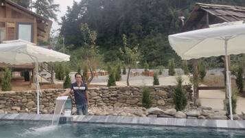 Zona de piscina amb un mur de pedra, una persona al costat de la piscina i un edifici de fusta al fons.