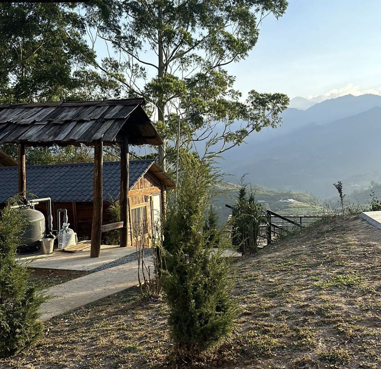 Una cabaña rústica con techo de metal, rodeada de árboles y una vista a montañas distantes.