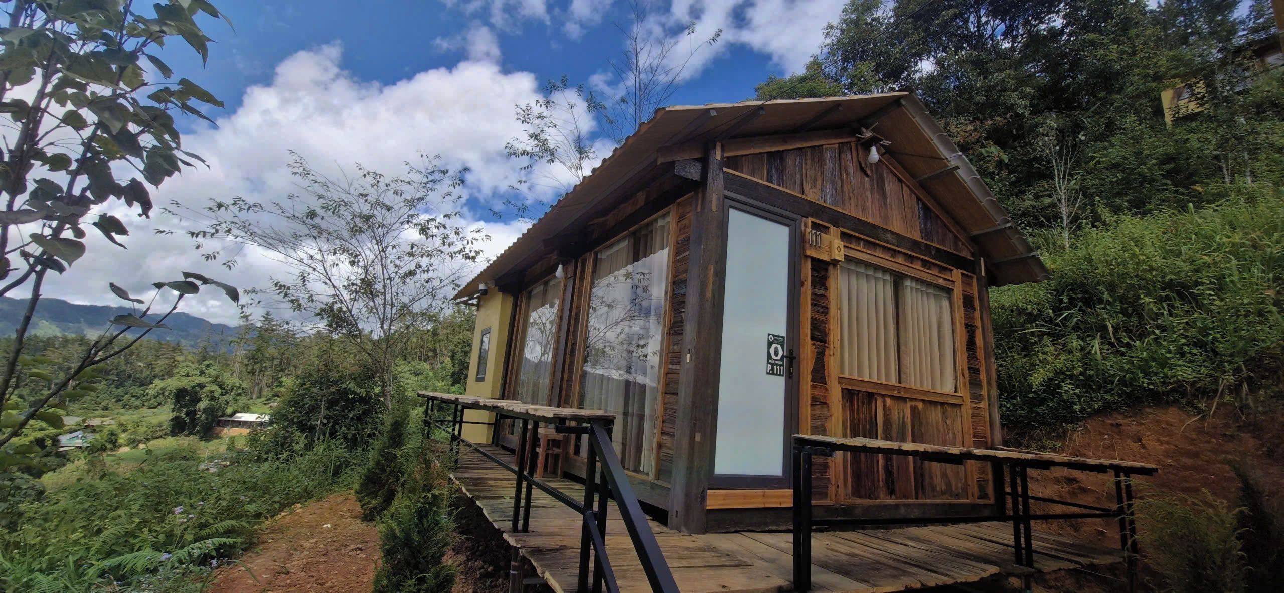 A wooden cabin with a balcony, surrounded by lush greenery and mountains in the background.