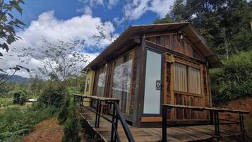 A wooden cabin with a balcony, surrounded by lush greenery and mountains in the background.